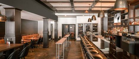 A bar area with long tables and stools, a bar counter with bottles, and a ceiling with exposed beams and pendant lights.