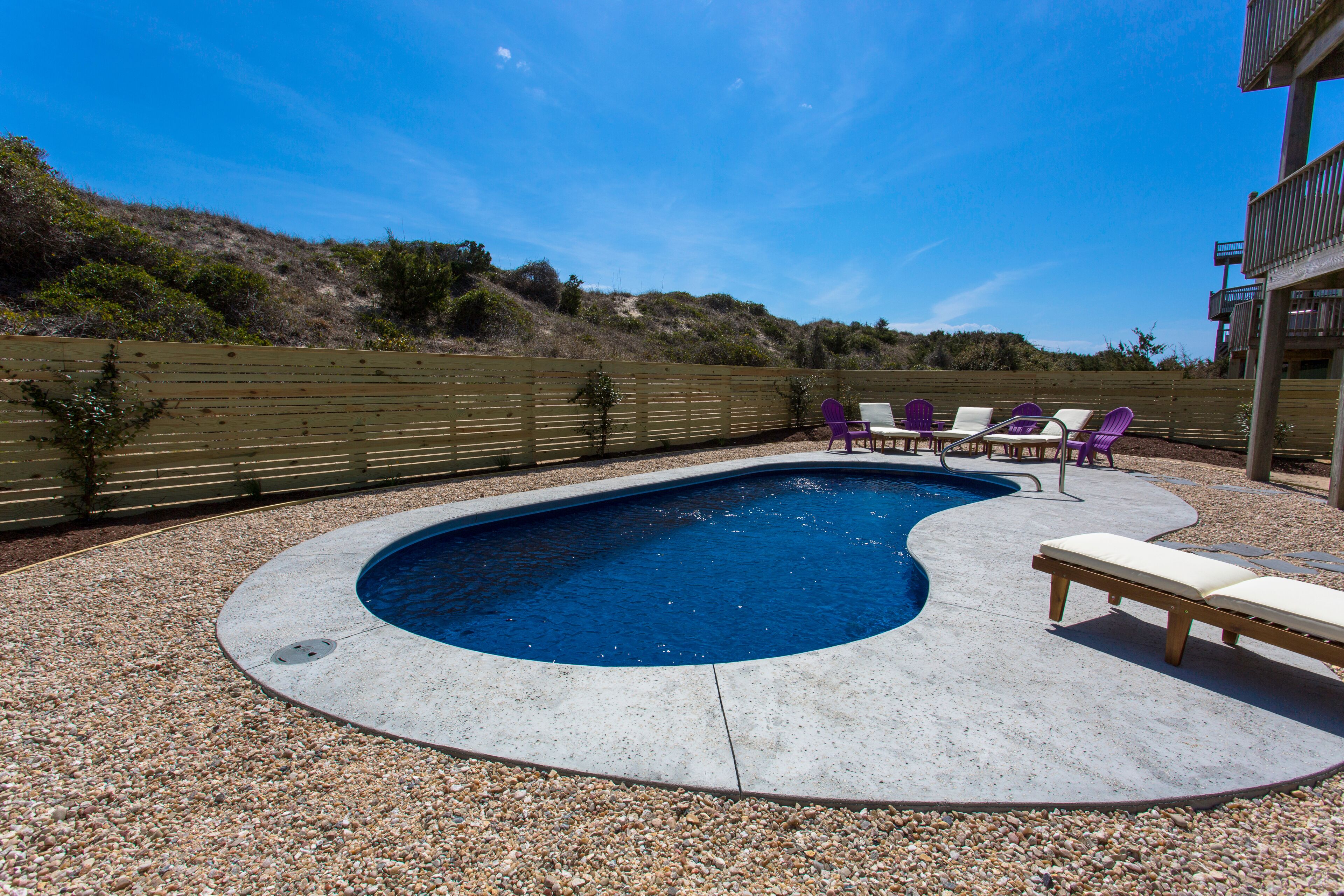An outdoor swimming pool with a pebble deck, surrounded by a wooden fence and a building with a balcony.