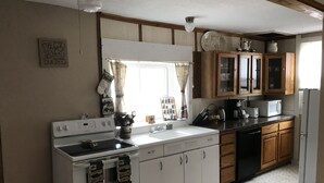 A compact kitchen with white appliances, wooden cabinets, and a window with curtains. - Vintage Cabin with Hot Tub, Convenient in-Town Location & Mountain Views! #3511 (Estes Park)