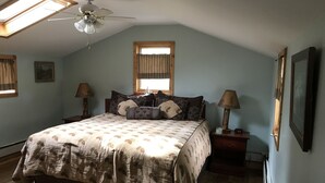 A bedroom with a sloped ceiling, a bed with patterned bedding, two bedside lamps, a ceiling fan, and a window with curtains. - Vintage Cabin with Hot Tub, Convenient in-Town Location & Mountain Views! #3511 (Estes Park)