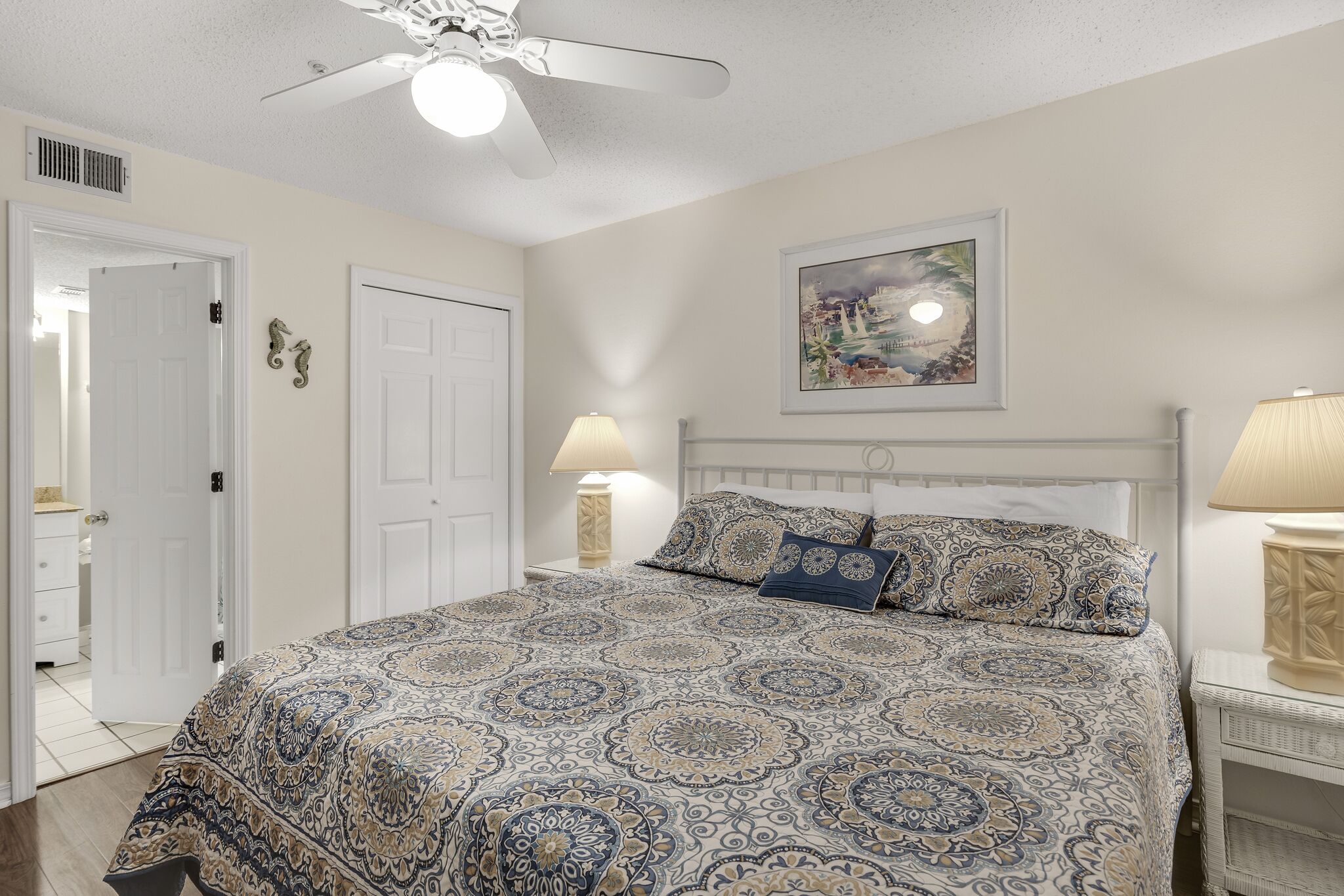 A bedroom with a patterned bedspread, a ceiling fan, a framed painting, and a bedside lamp.