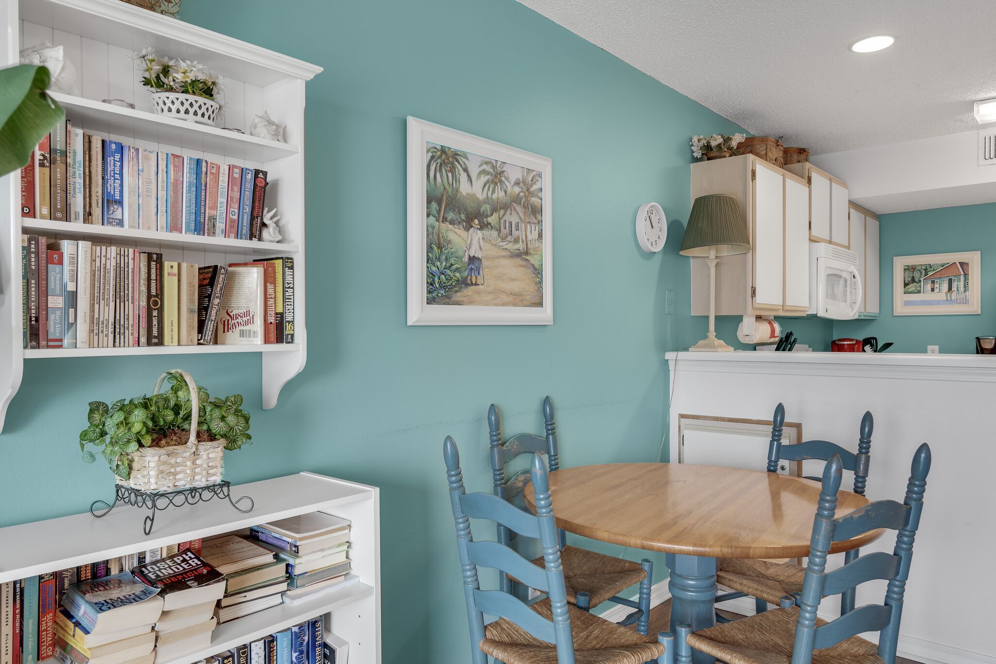 Un comedor con mesa de madera y sillas azules, una estantería con libros y un reloj fijado a la pared.