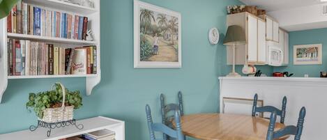 Un comedor con mesa de madera y sillas azules, una estantería con libros y un reloj fijado a la pared.