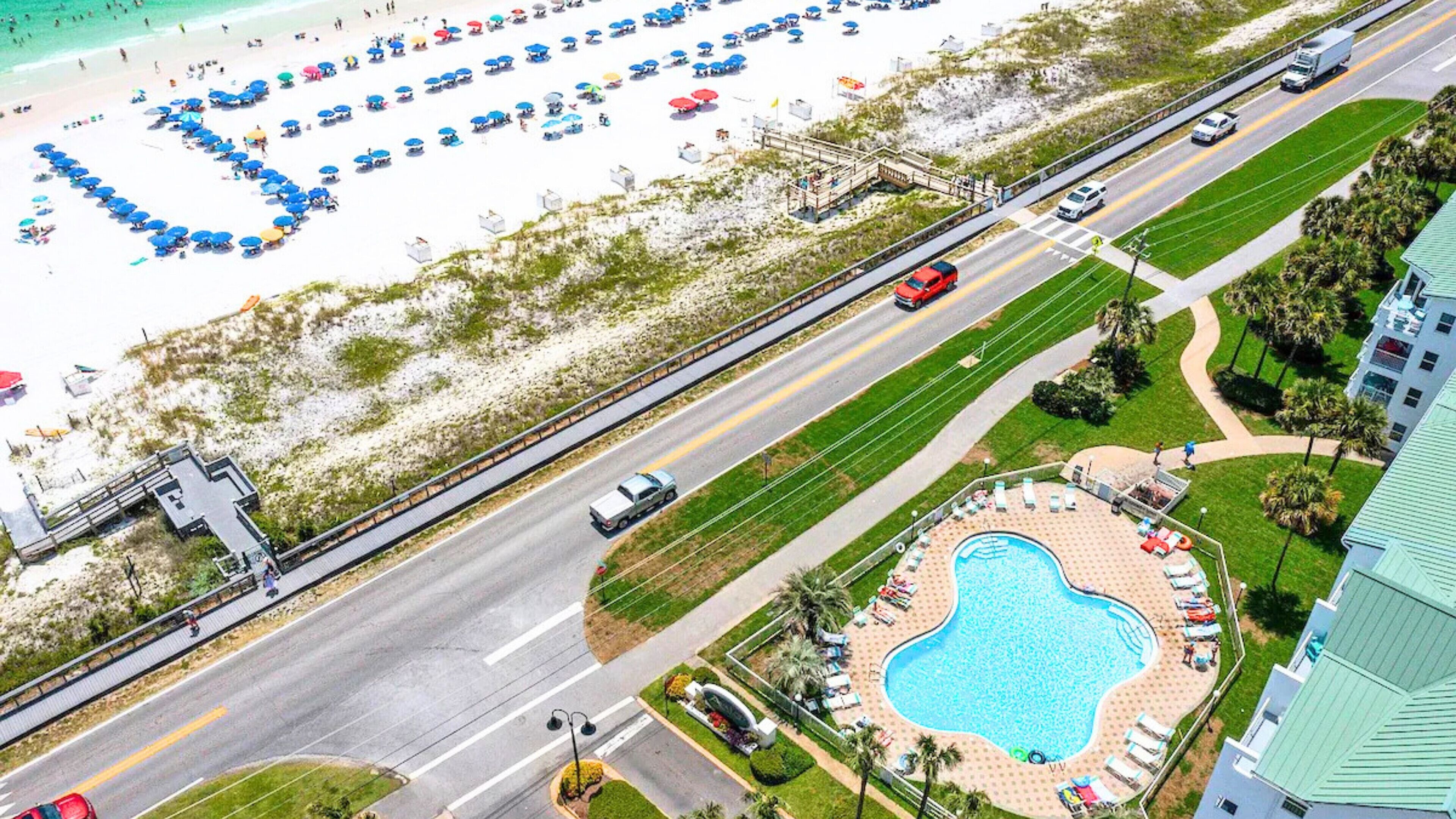 Vista aérea de un resort frente a la playa, con piscina, sombrillas y una carretera con vehículos.