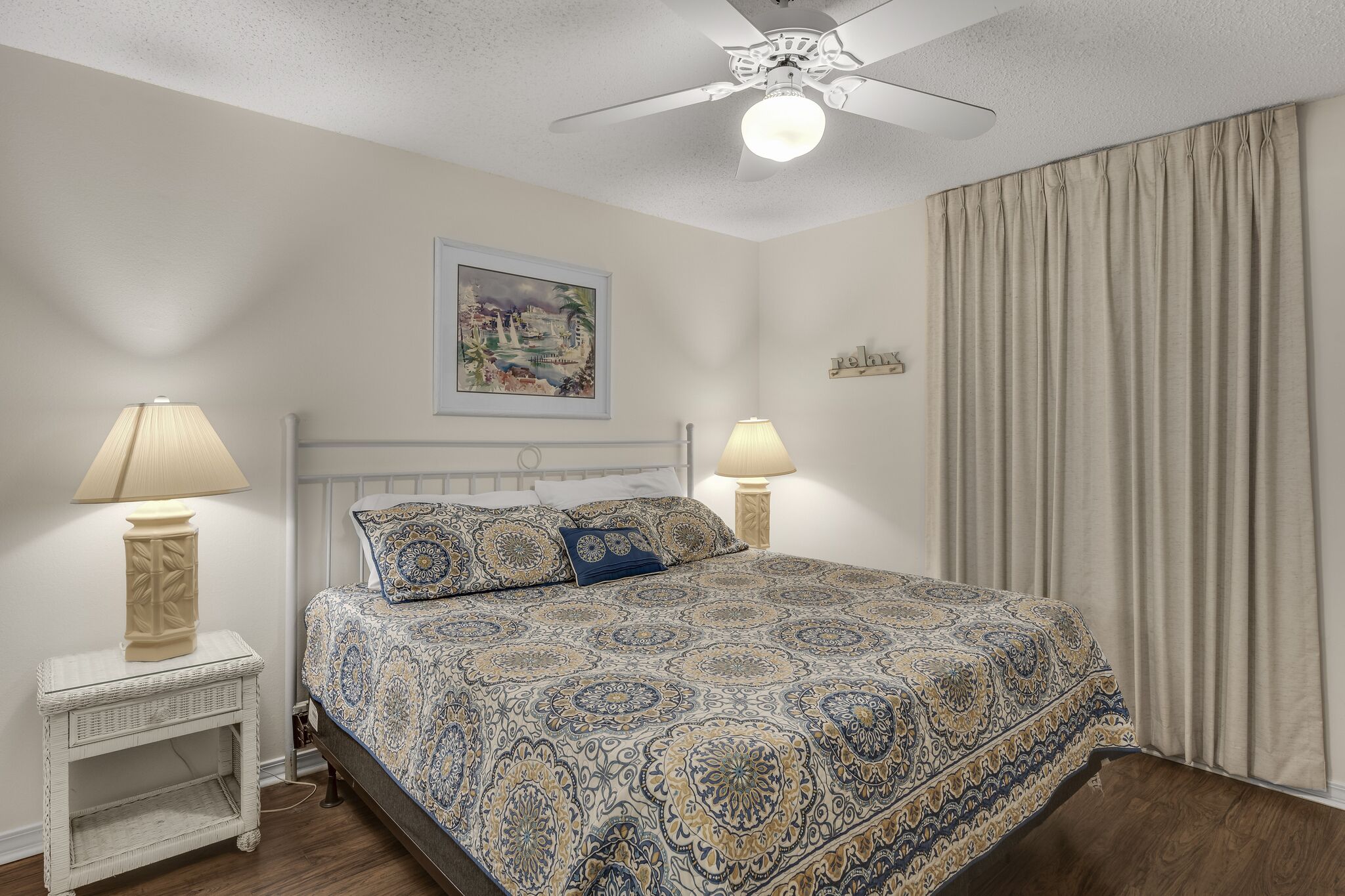 Una camera da letto con una coperta dal disegno decorativo, due lampade da comodino, una ventola a soffitto, un quadro appeso al muro e un'ampia finestra con tende.