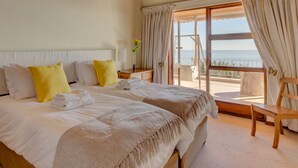 Un lit double avec des oreillers blancs et jaunes, une table de chevet en bois avec une lampe, et une vue sur la mer par une grande fenêtre ornée de rideaux.