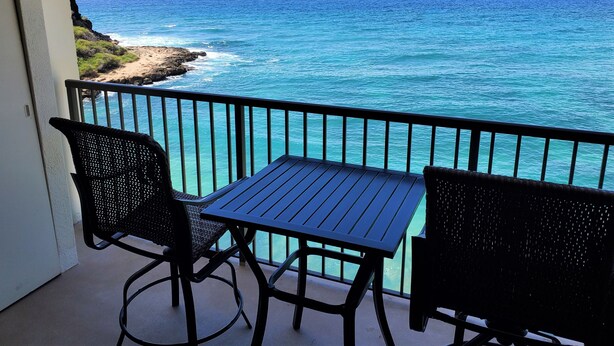 Varanda com mesa e duas cadeiras, vista para a costa, oceano e um veleiro.