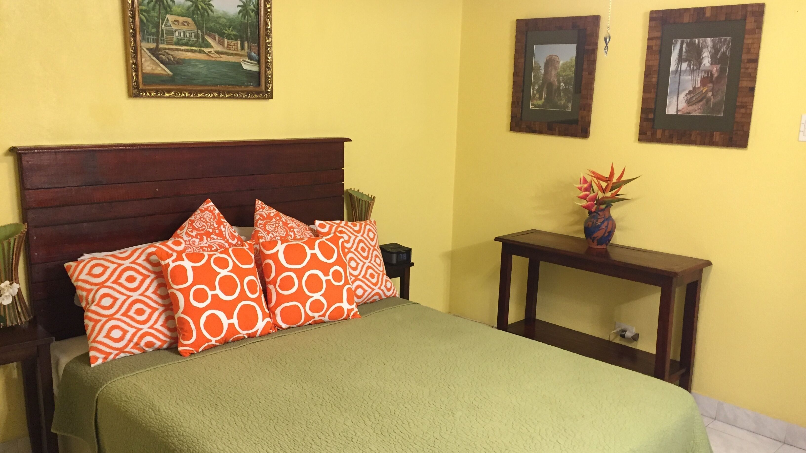 Una cama con colcha verde, almohadones estampados anaranjados, cabecera de madera y mesita de noche con un florero.