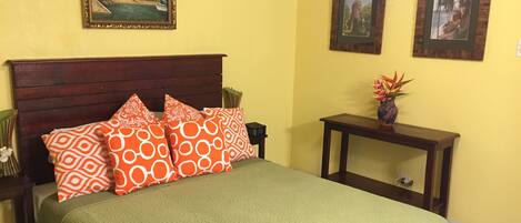 Una cama con colcha verde, almohadones estampados anaranjados, cabecera de madera y mesita de noche con un florero.