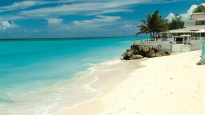 Una playa con agua turquesa cristalina, una orilla arenosa y un edificio con techo blanco y palmeras.