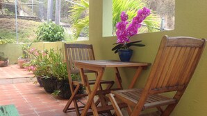 Une table et des chaises en bois sont disposées sur une terrasse carrelée, agrémentée de plantes en pot et d’une fenêtre ornée de fleurs violettes.