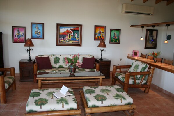 A living room with a sofa, two armchairs, a coffee table, and a small table with a lamp. There are four framed paintings on the wall.
