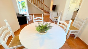 A round white table with a potted plant in the center, surrounded by white chairs with woven seats. A staircase with wooden railings and a white cabinet with a television are visible in the background. - 0681 Villa Molins Libra (Denia)