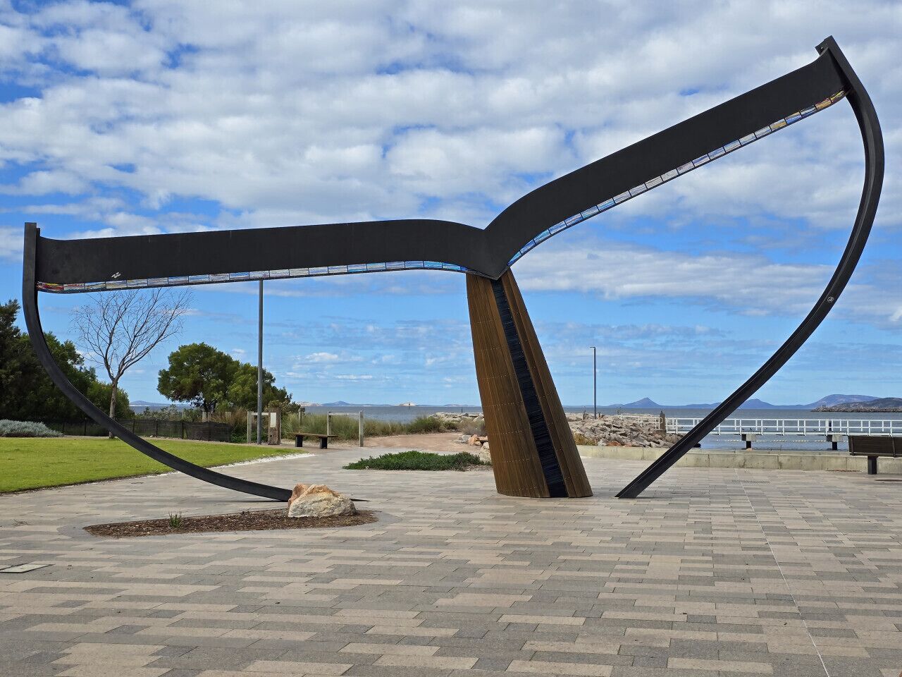 Une sculpture en plein air, installée dans un espace public, avec un ciel dégagé et des montagnes à l’horizon.