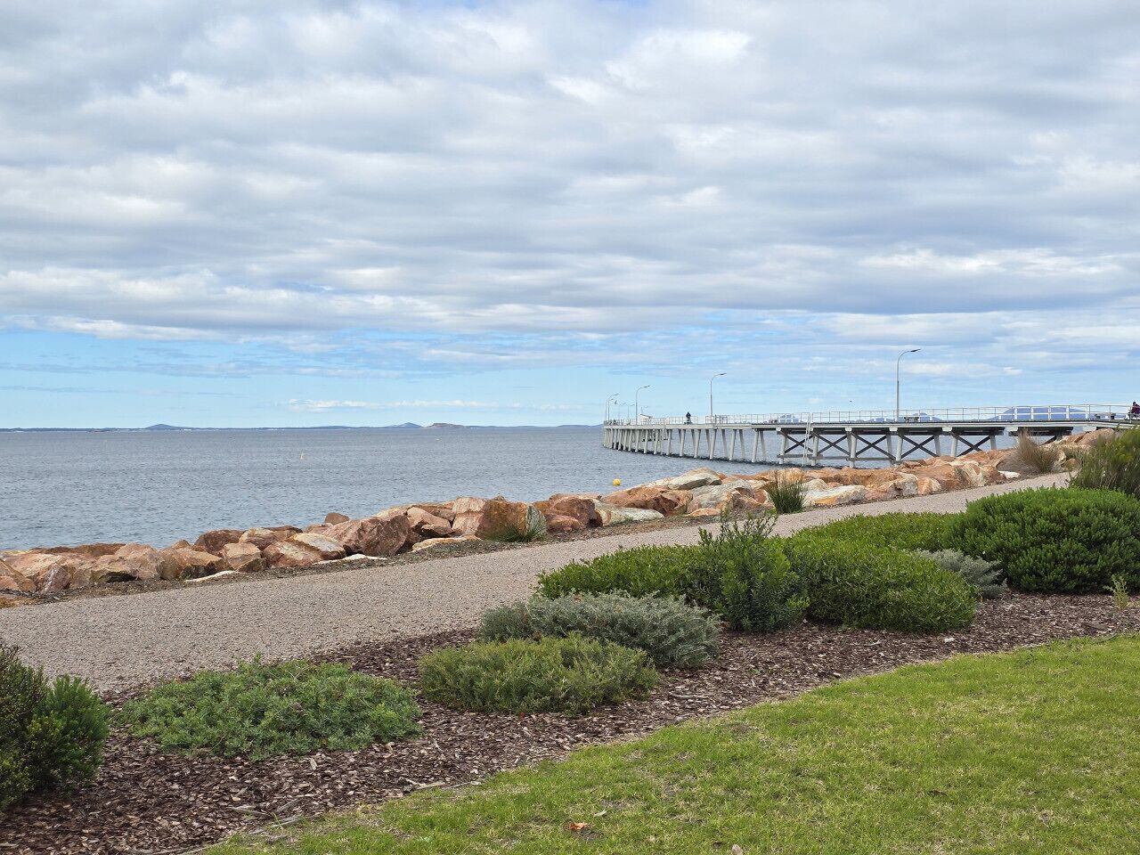 A coastal landscape with a rocky shoreline, a grassy area, and a pier extending into the water.