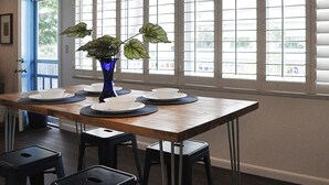 A dining area with a wooden table set for four, a blue vase with a plant, and a sign reading "BEACH HOUSE" above the windows.