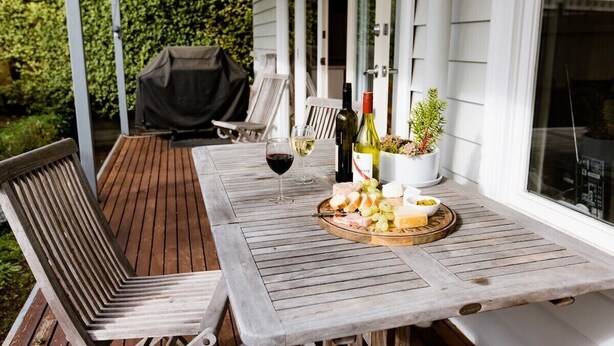 A wooden deck with a table set for a meal, including a cheese platter and wine glasses. - Luxury Accessible Garden Retreat Near Beaches, Golf & Wineries (Point Lonsdale)
