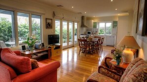 A spacious living area with a red sofa, a patterned armchair, and a dining table with chairs. A television and speakers are on a wooden stand. The kitchen is visible in the background with a microwave and cabinets. - Luxury Accessible Garden Retreat Near Beaches, Golf & Wineries (Point Lonsdale)