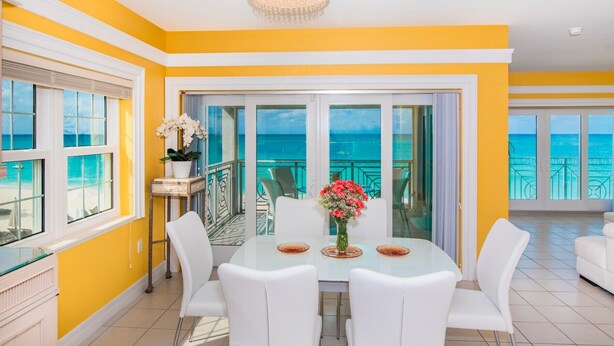 Una habitación luminosa de color amarillo con una mesa de vidrio y sillas blancas, un candelabro y vista al océano.