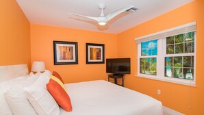 Habitación de hotel con una pared naranja, una cama con ropa blanca, dos cuadros enmarcados, un ventilador de techo, un televisor y una ventana con vistas a palmeras.