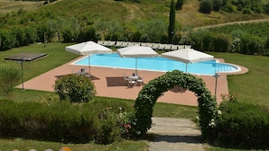 A swimming pool with white umbrellas, surrounded by a garden with a wooden pathway leading to a seating area.