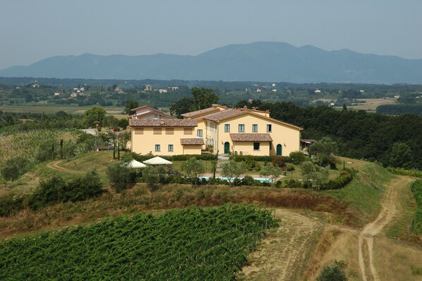 Une grande villa jaune dotée d’une piscine, entourée de vignes et de collines.