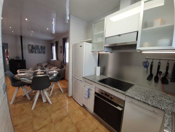 A compact kitchen with a built-in oven, a stovetop, and a refrigerator. There is a dining area with a table and chairs, and a sofa in the background. - House in perfect condition, quiet area 750m from the beach, close to supermarkets. (L'Escala)
