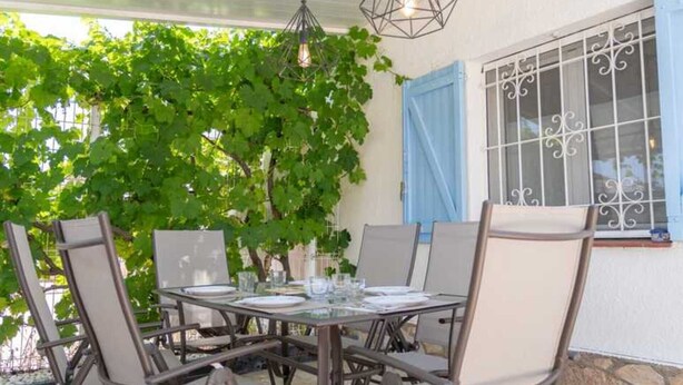 Un espace de restauration en plein air, avec une table et des chaises, entouré de verdure et agrémenté d’une fenêtre ornée de barreaux décoratifs.