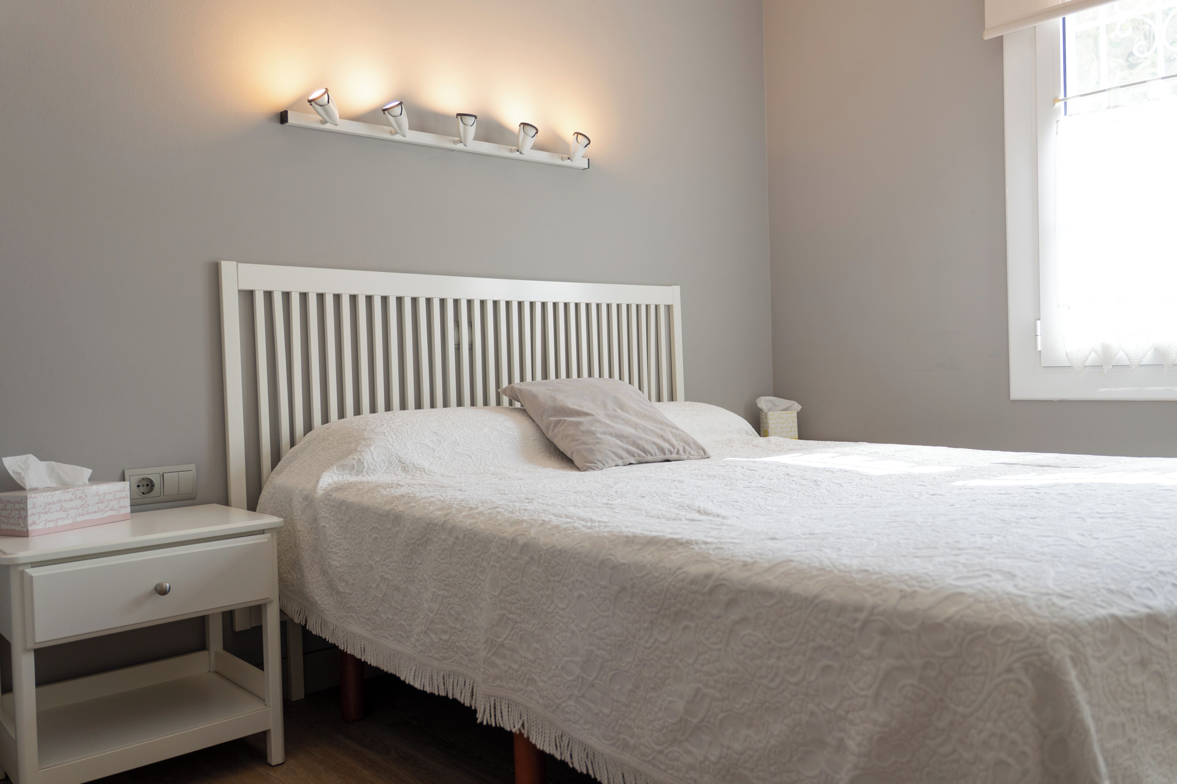 A neatly made bed with a white headboard, a bedside table with a lamp, and a wall-mounted light fixture.