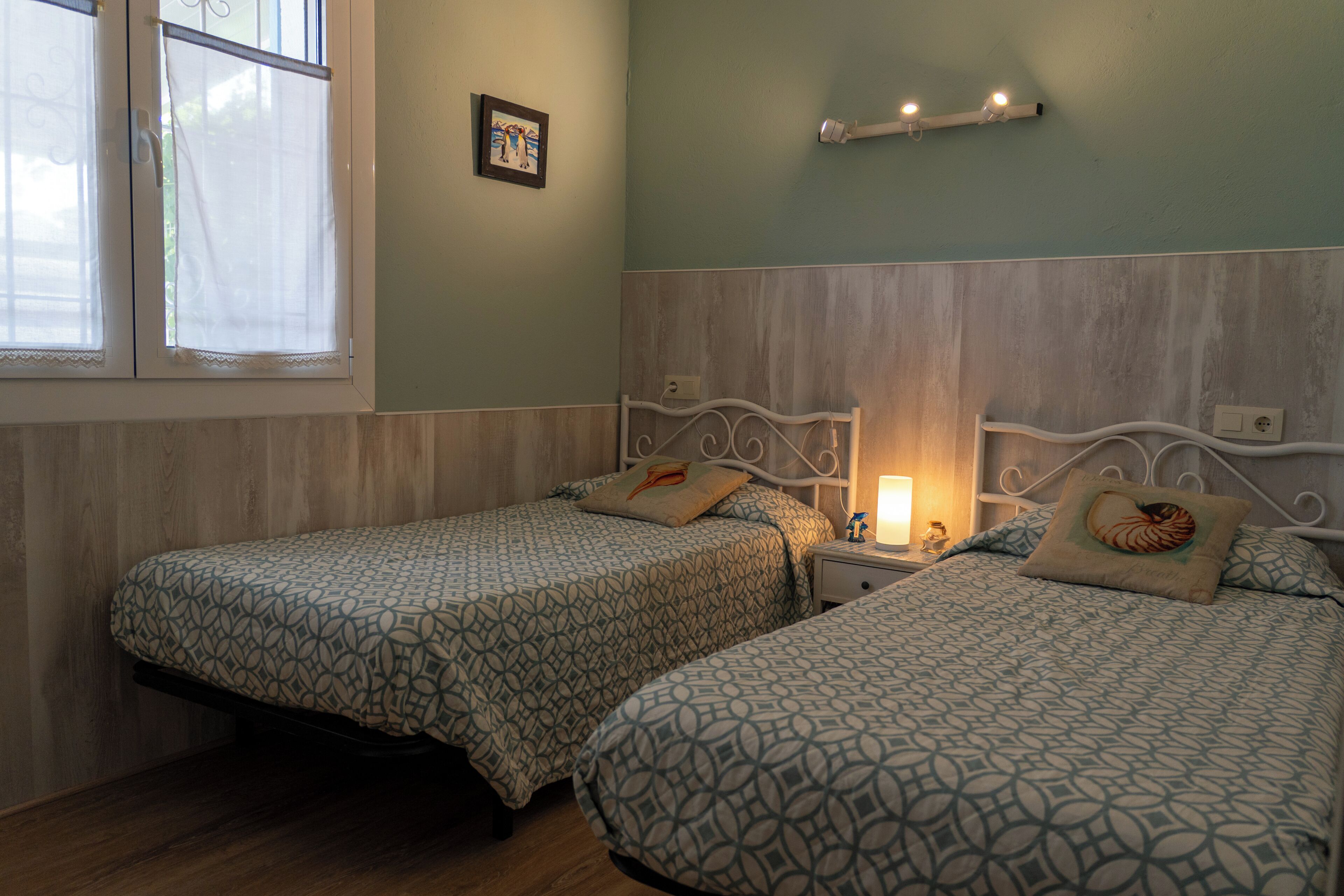 A room with two beds, a nightstand with a lamp, a wall-mounted shelf, and a window with curtains.