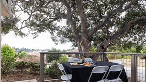 An outdoor dining area with a table set for four, surrounded by white chairs, under a large tree.