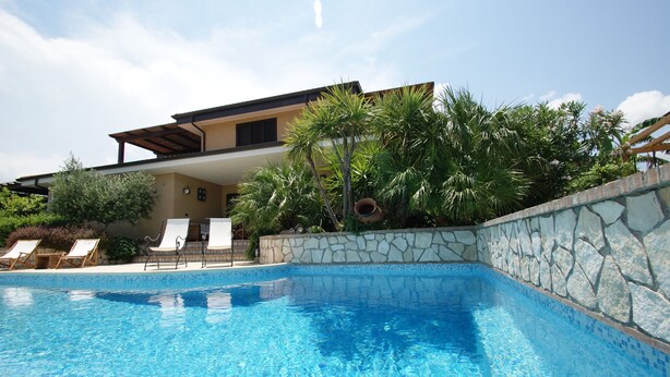 A swimming pool with lounge chairs, a stone wall, and a modern house with a pergola. - beautiful detached villa with private pool wi-fi (Briatico)