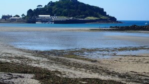 Un paysage côtier avec un château perché sur une colline, une plage de sable et une eau calme.