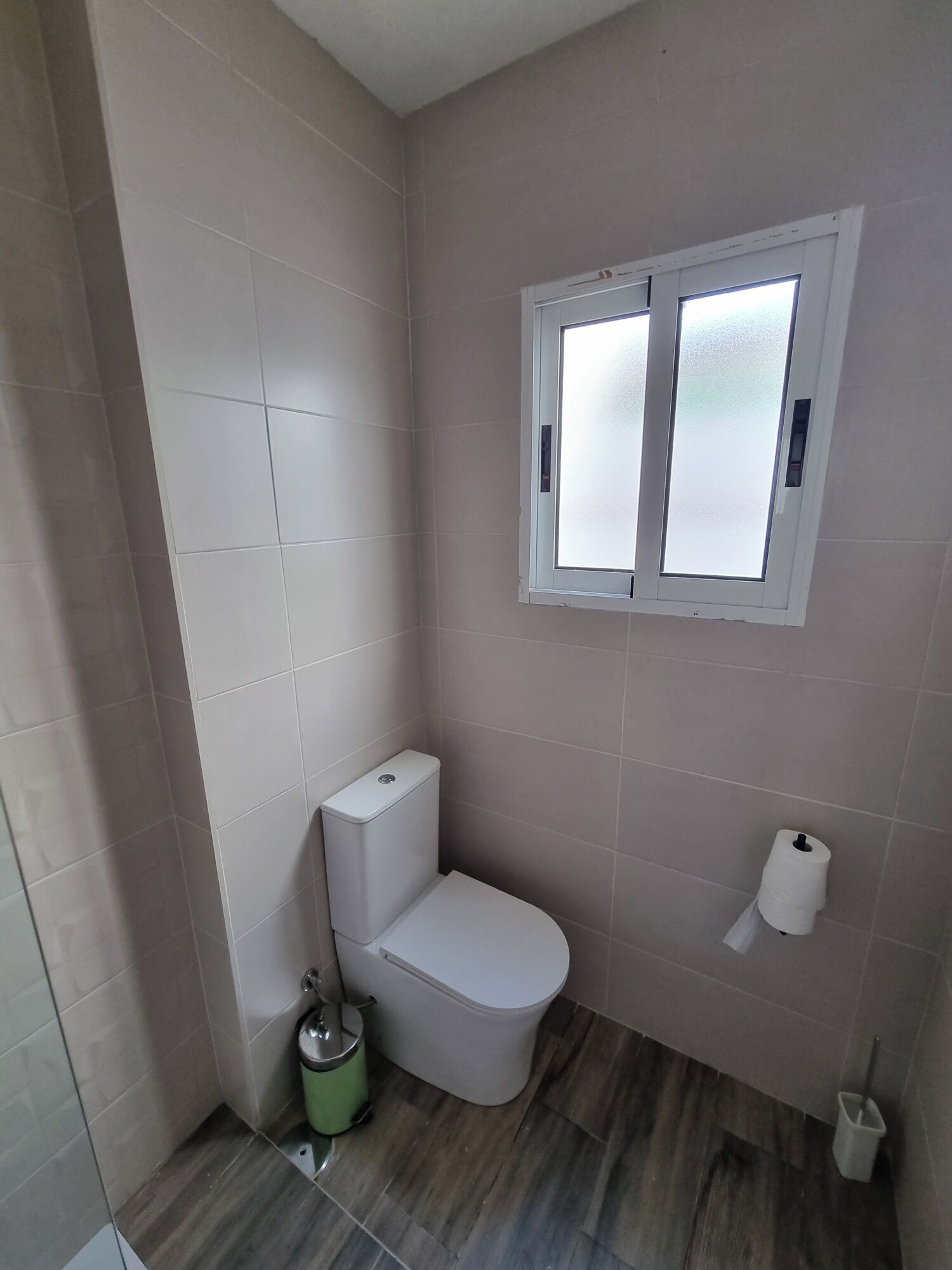 Une petite salle de bain propre, avec des toilettes, une poubelle verte, un porte-rouleau de papier toilette blanc et une fenêtre à carreaux dépolis.