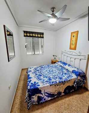 Un dormitorio con cama, ventilador de techo, ventana con persianas, mesita de noche con un florero y un cuadro colgado en la pared.