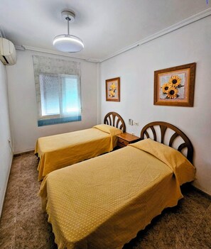 Quarto com duas camas, janela com persianas, luminária no teto e quadros na parede.