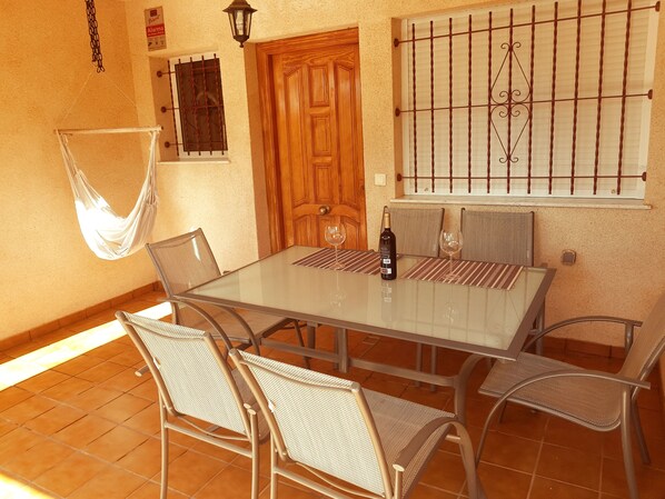 A dining area with a glass table, wicker chairs, a wooden door, a barred window, and a hanging hammock.