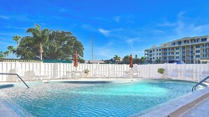 Ein Swimmingpool mit Liegestühlen und Sonnenschirmen, umgeben von einem weißen Zaun, unter blauem Himmel.