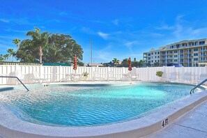 Ein Swimmingpool mit Liegestühlen und Sonnenschirmen, umgeben von einem weißen Zaun, unter blauem Himmel.