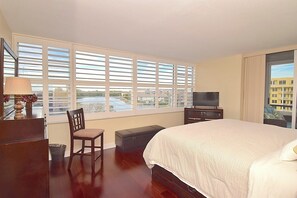 A hotel room with a large bed, a television, a desk with a lamp, a chair, and a view of a body of water through the windows.