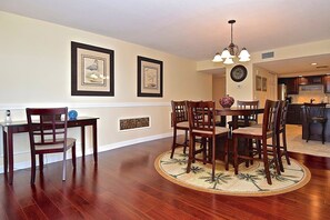 A dining area with a round table and chairs, a chandelier, framed artwork on the wall, and a kitchen in the background.