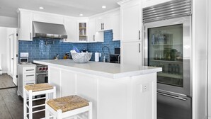Una cocina moderna con armarios blancos, salpicadero azul, extractor de acero inoxidable, horno empotrado, refrigerador y una isla grande con taburetes de bar.