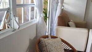 Ett mysigt vardagsrum med en flätstol, ett litet bord med dekorativa föremål, en vit soffa och ett fönster med vita gardiner.