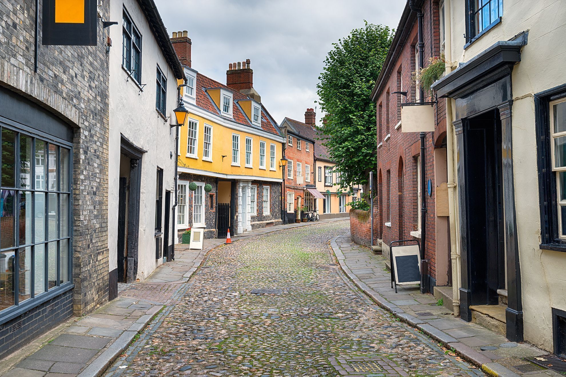 A cobblestone street lined with historic buildings, including a yellow building with a black awning and a brick building with a black door.