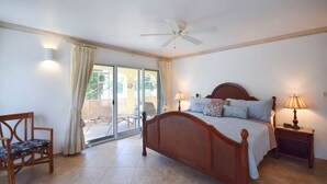 Un dormitorio con cama, dos mesitas de noche con lámparas, una silla, un ventilador de techo y un ventanal corredero que da a un espacio exterior.