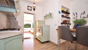 A kitchen with a stove, oven, and a dining area with chairs. There is a bookshelf and a potted plant. - Villa with 3 bedrooms and 2 bathrooms in residence (Aglientu)