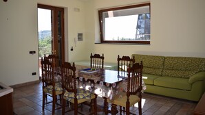 A dining area with a glass-top table surrounded by wooden chairs, a green sofa, and a window with a view of greenery and buildings.
