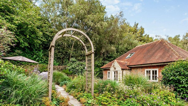 A rustic house with a red-tiled roof, surrounded by lush greenery and a wooden archway.