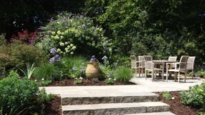 A garden patio with stone steps leading to a wooden table and chairs, surrounded by lush greenery and flowering plants.