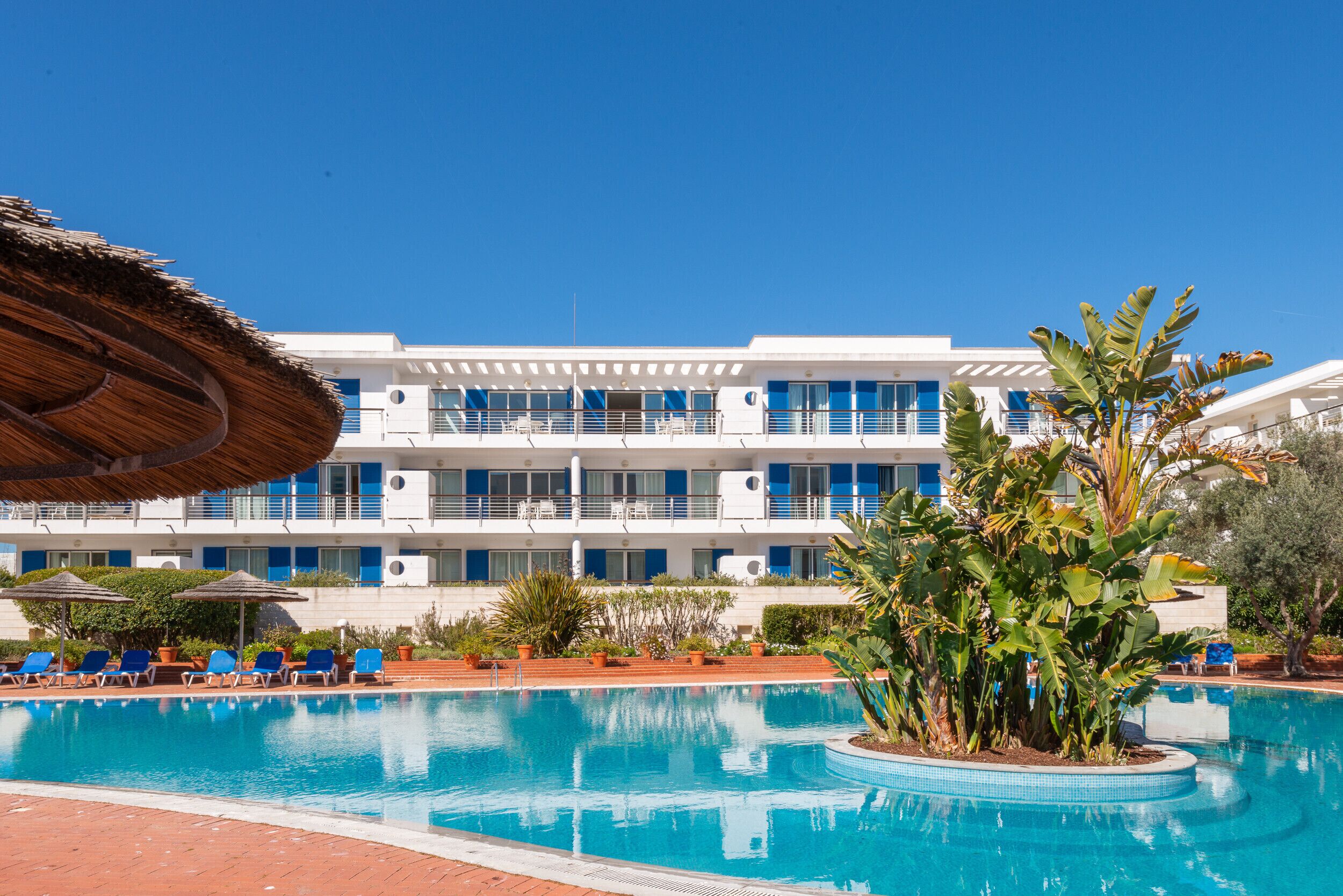 A hotel with a swimming pool, blue chairs, and a thatched umbrella.